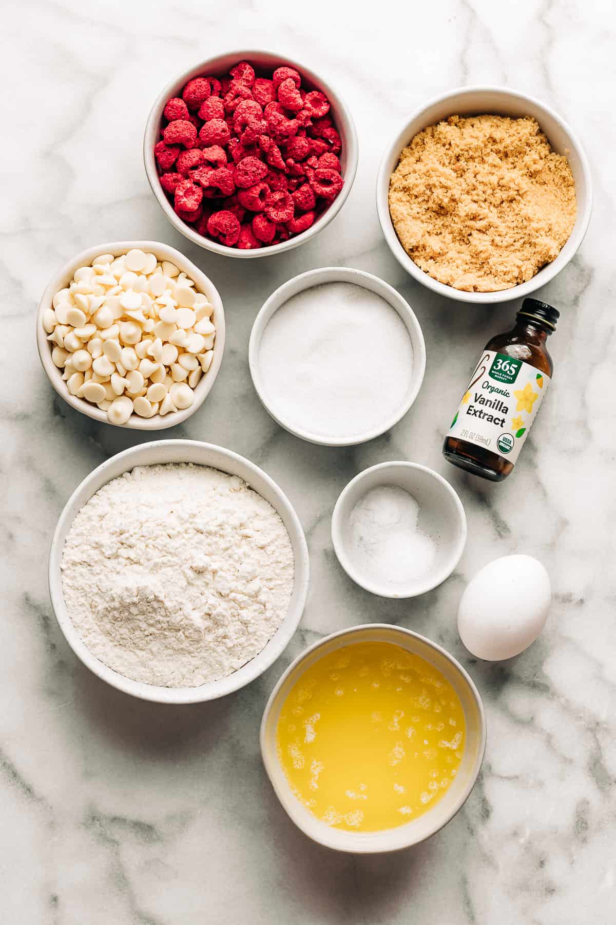 freeze dried raspberries, light brown sugar, white chocolate chips, sugar, flour, baking soda, salt, vanilla extract, egg, and melted butter on a marble surface.