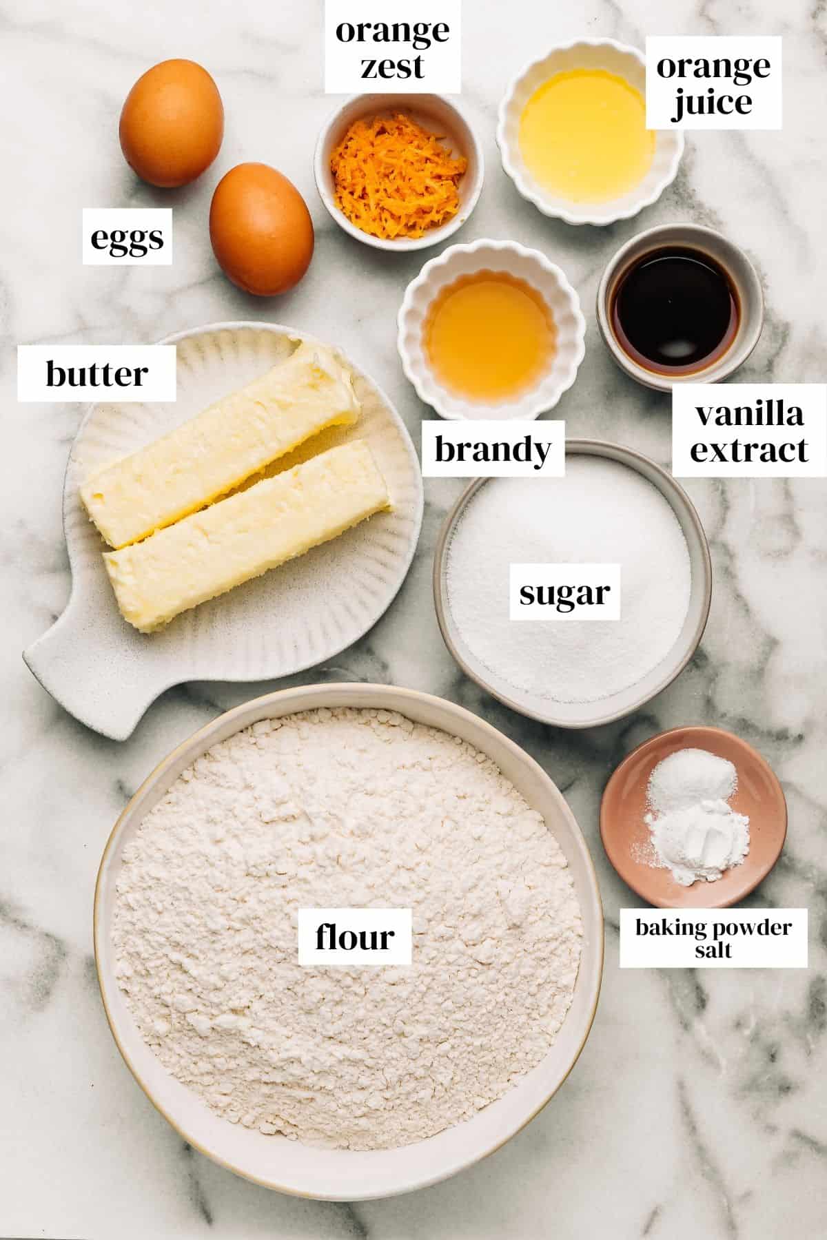 eggs, orange zest, orange juice, brandy, vanilla extract, sugar, butter, baking powder, salt, and flour on a marble surface.