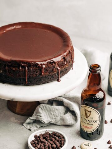 chocolate cheesecake with ganache on a marble cake stand next to a guinness beer bottle and chocolate chips.