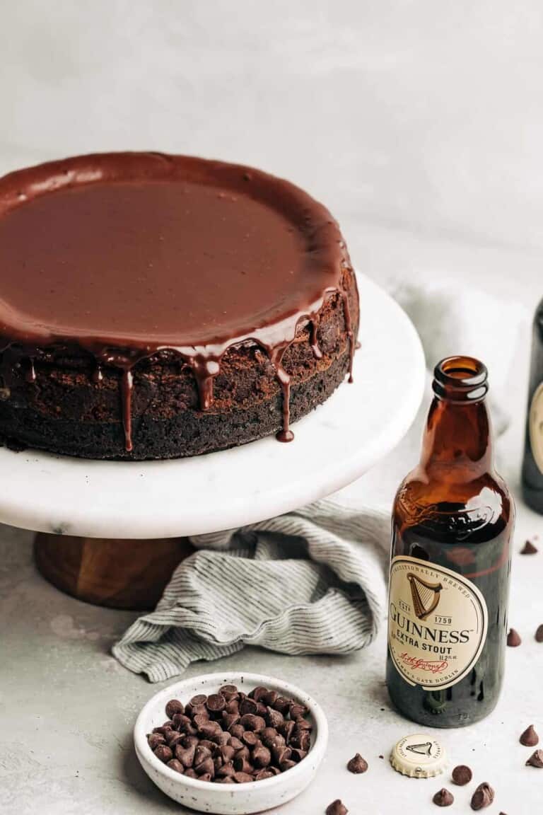 chocolate cheesecake with ganache on a marble cake stand next to a guinness beer bottle and chocolate chips.