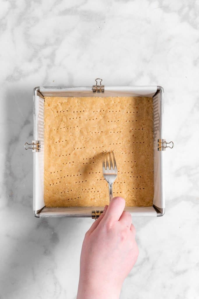 shortbread crust in a baking pan being pricked with a fork