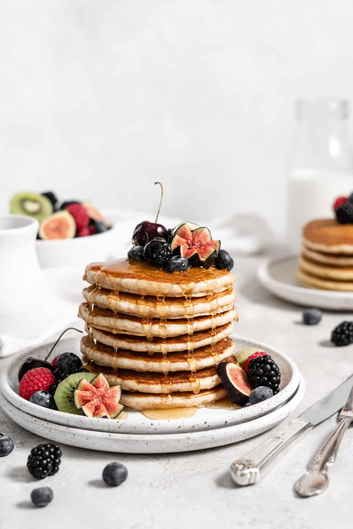 stack of pancakes on a plate with sliced fruit, berries, and maple syrup.