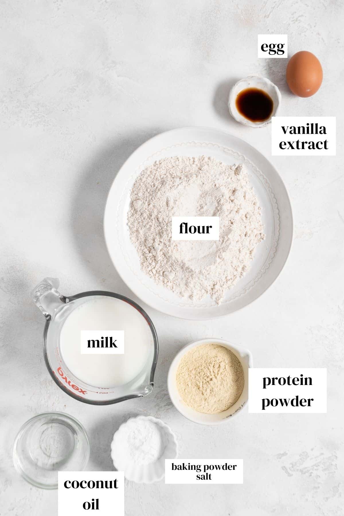 egg, vanilla extract, flour, milk, protein powder, baking powder, salt, and melted coconut oil on a light surface.