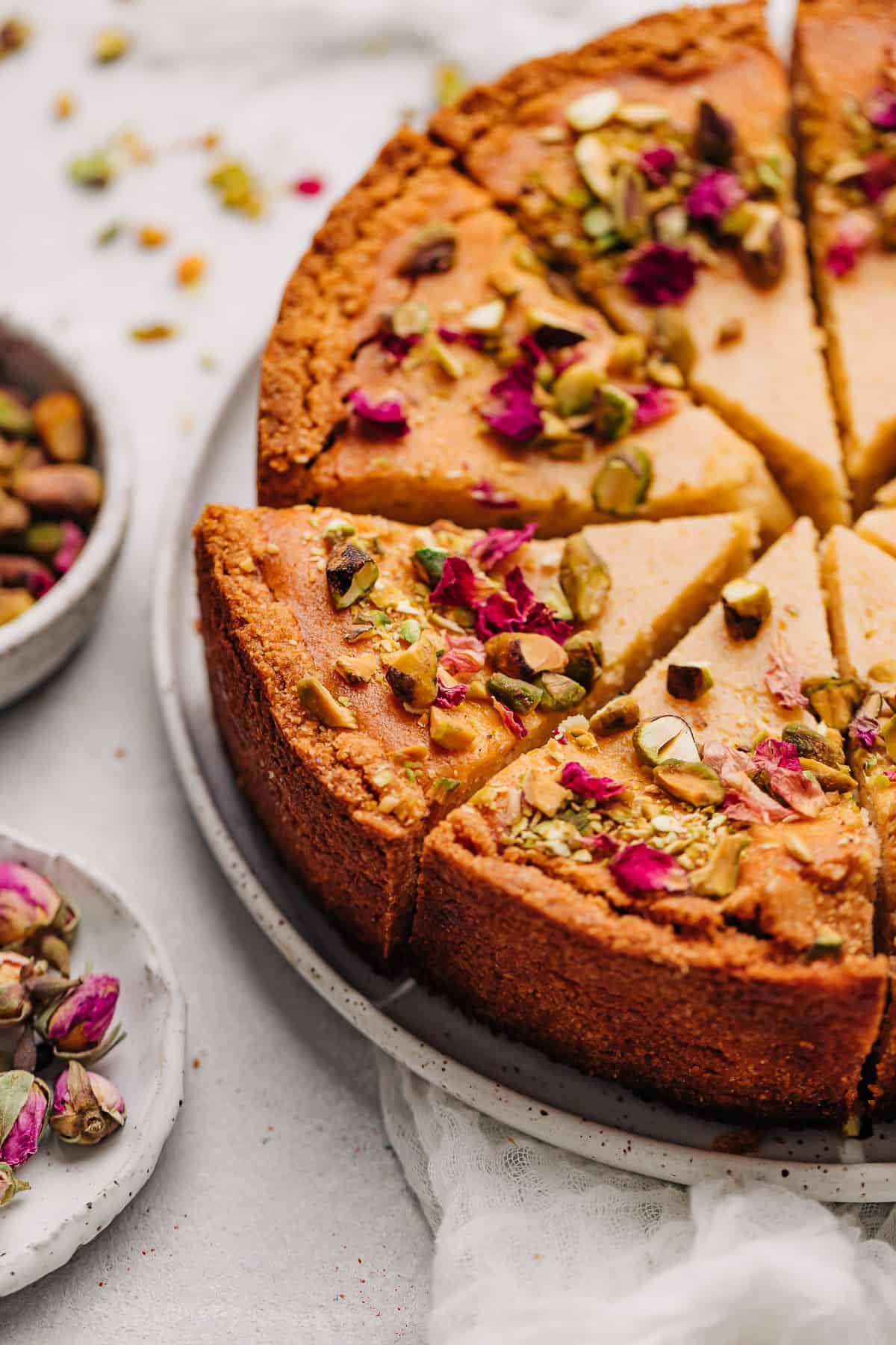 Slices of Persian Love Cake on a plate with small bowls of chopped pistachios and dried rose petals.