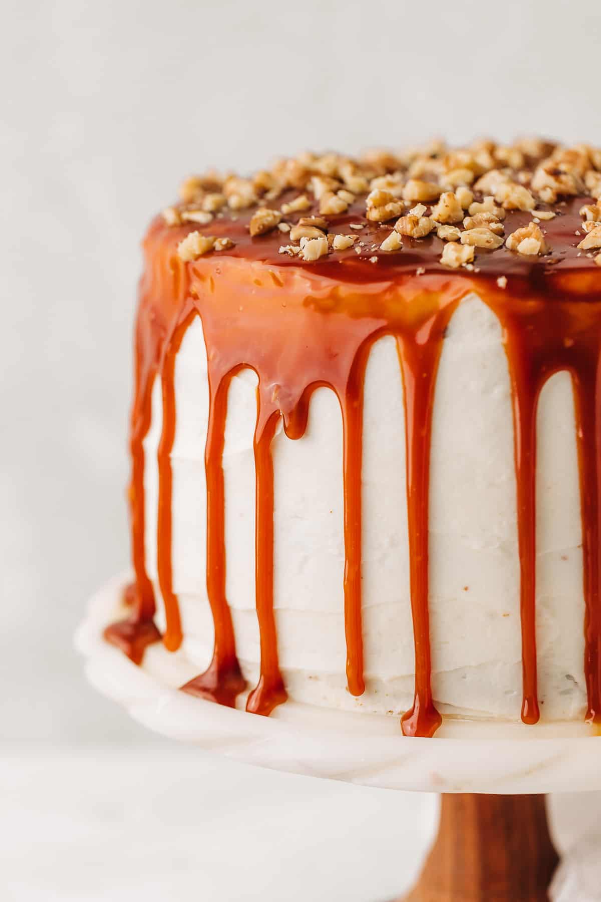 Carrot cake with cream cheese frosting topped with caramel sauce and chopped walnuts on a marble cake stand.