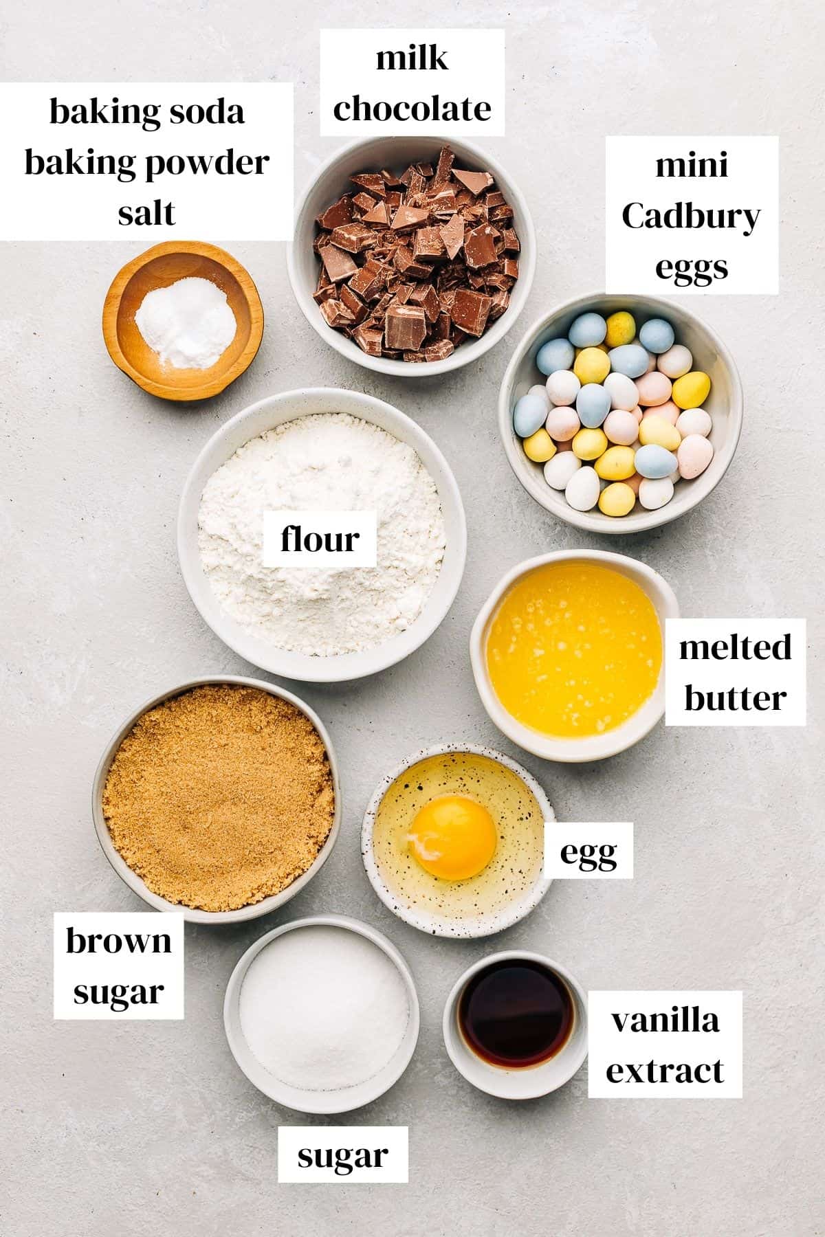 milk chocolate, cadbury mini eggs, melted butter, egg, vanilla extract, sugar, brown sugar, flour, baking powder, baking soda, salt on a gray surface.