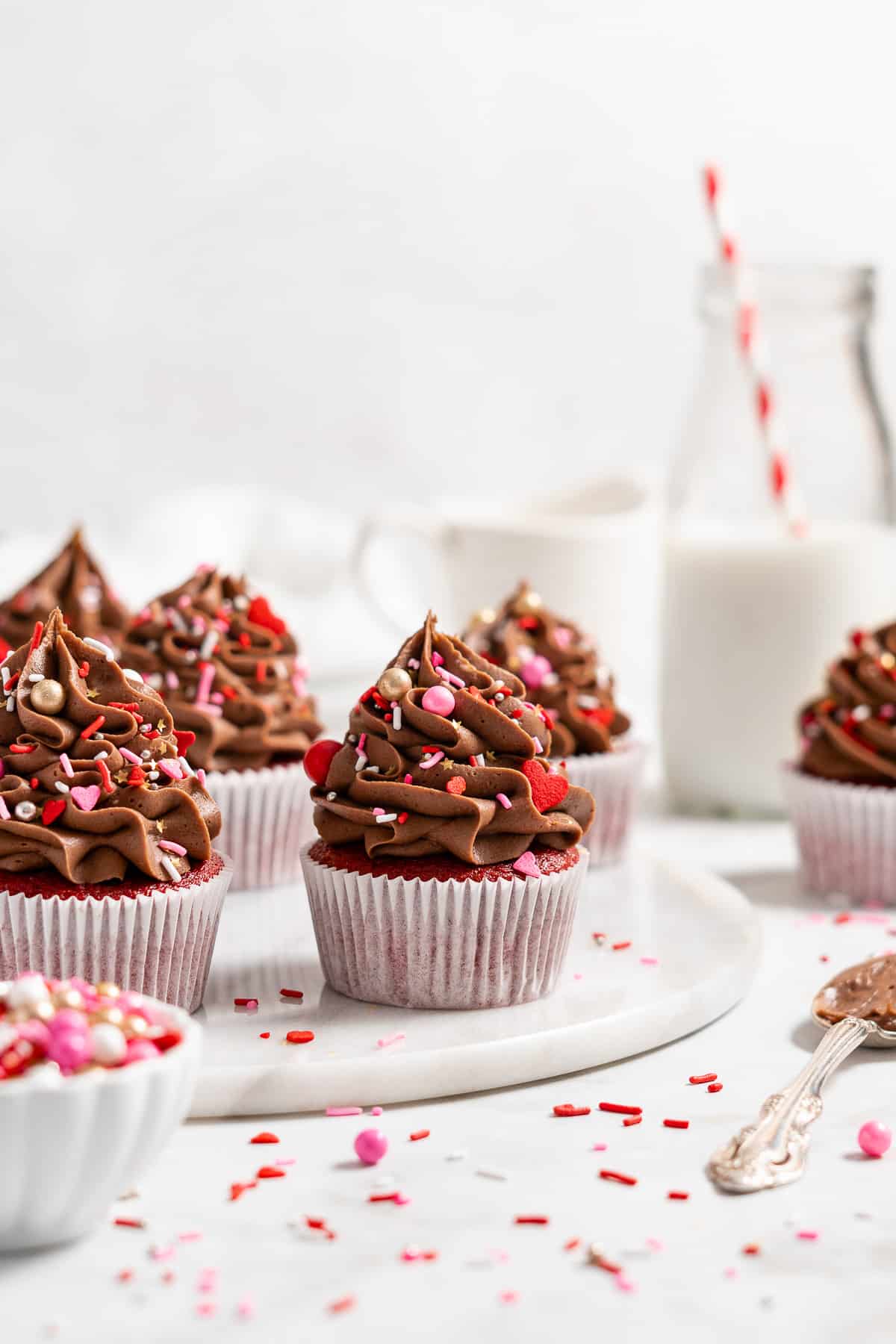 Red velvet cupcakes with chocolate frosting and pink and red sprinkles on a round marble slab.