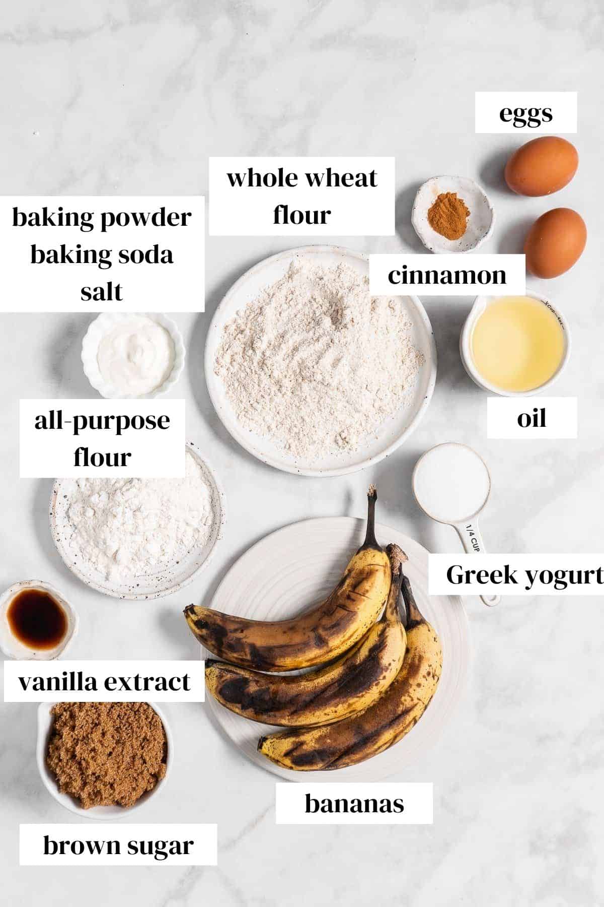 eggs, cinnamon, oil, yogurt, whole wheat flour, all purpose flour, bananas, vanilla, brown sugar on a marble surface.