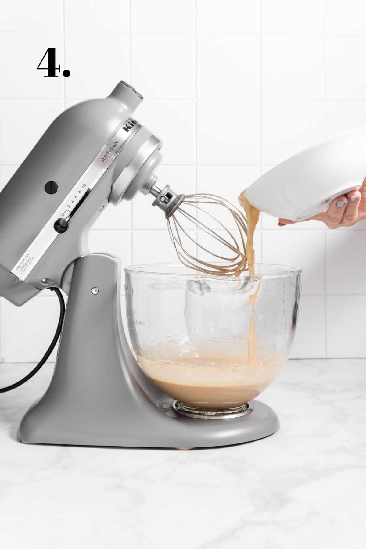 stand mixer with a glass bowl holding cake batter and mashed banana being poured in.