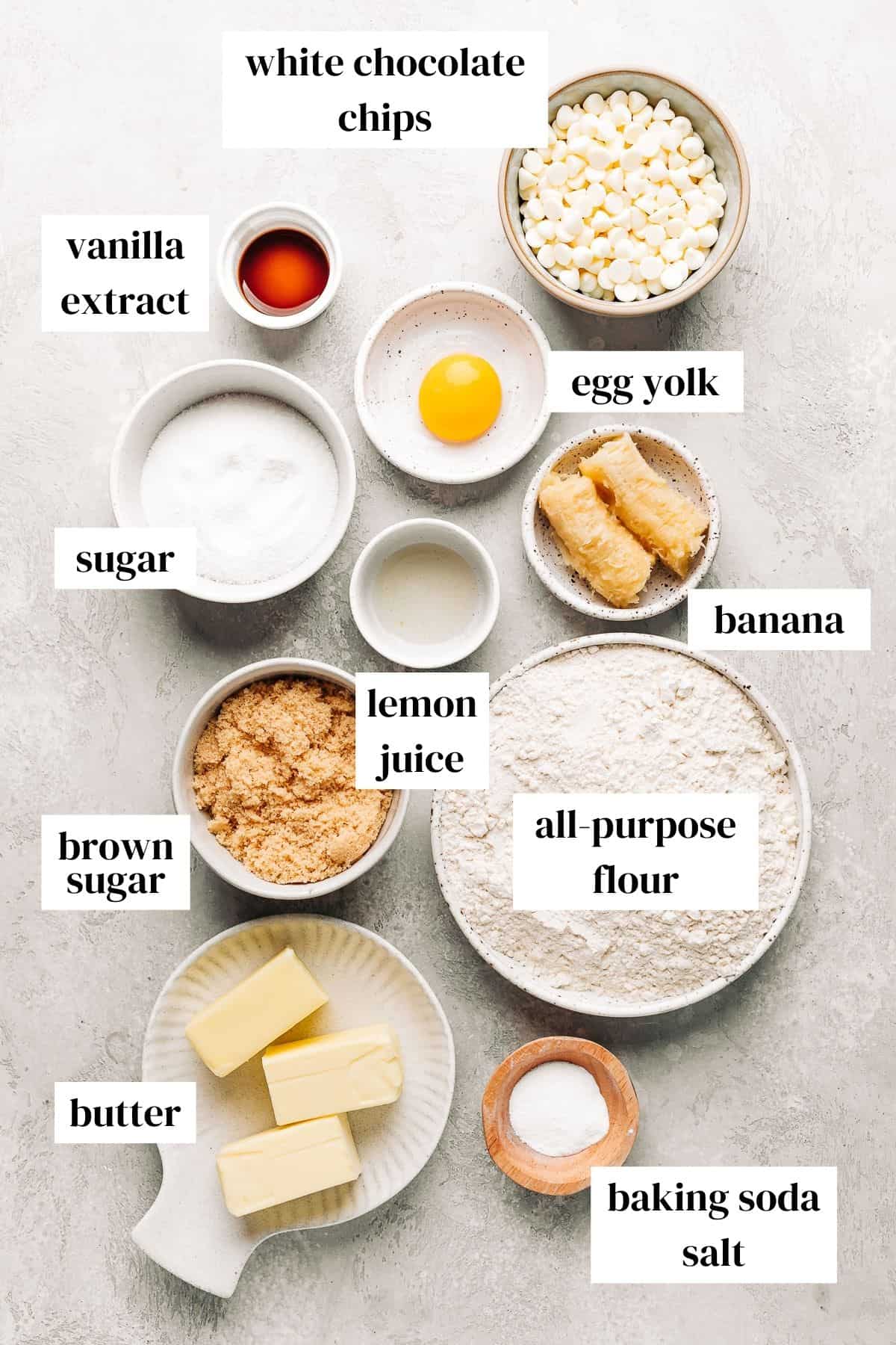 vanilla extract, white chocolate chips, egg yolk, banana, sugar, lemon juice, flour, brown sugar, butter, and salt on a grey background.