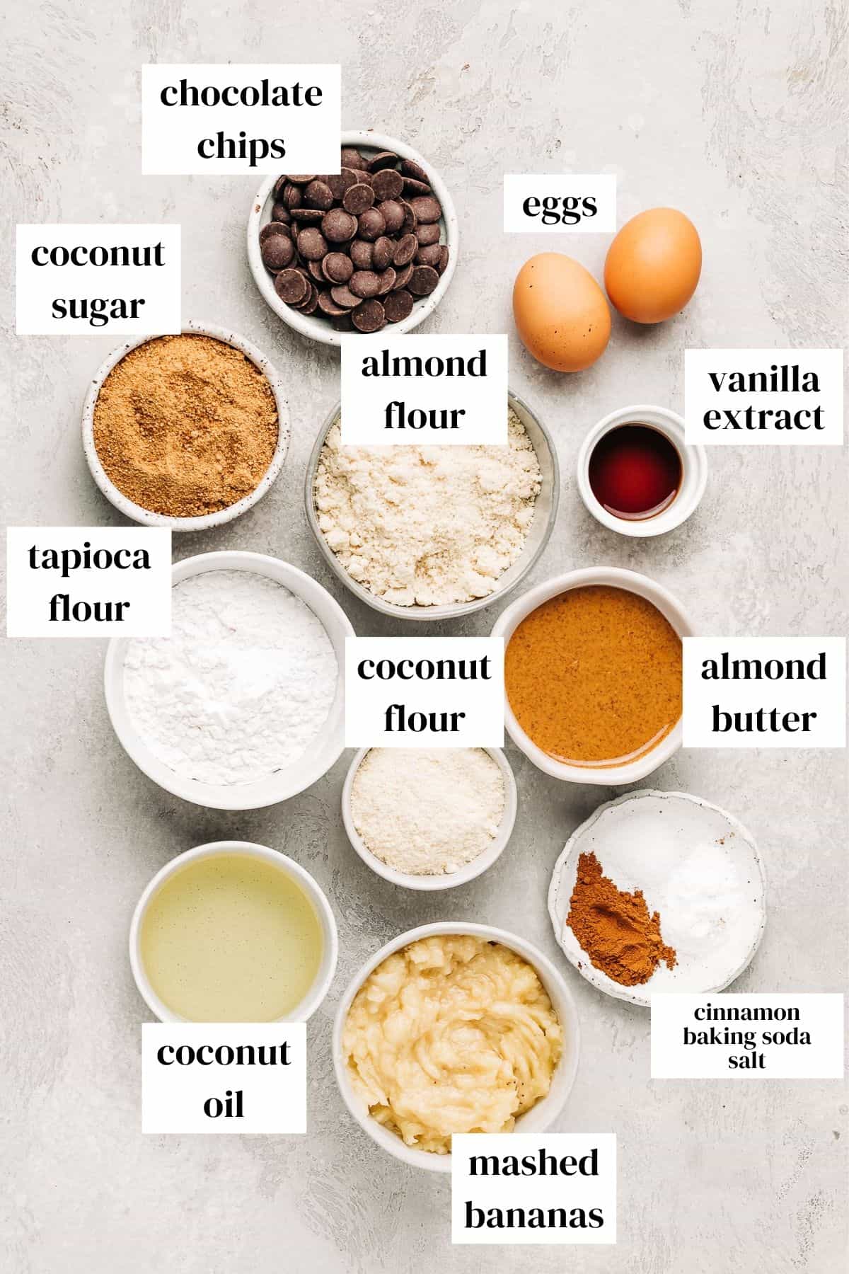eggs, chocolate chips, vanilla, coconut sugar, almond flour, tapioca flour, coconut flour, almond butter, coconut oil, bananas, cinnamon, baking soda, and salt on a gray surface.