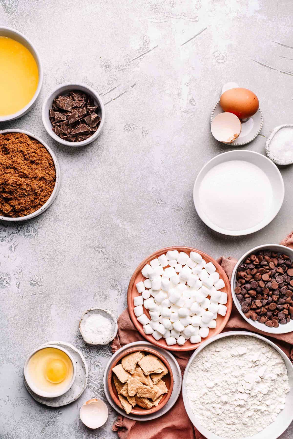 Melted butter, chocolate, brown sugar, eggs, sugar, chocolate chips, mini marshmallows, baking powder, flour, graham crackers in small bowls.