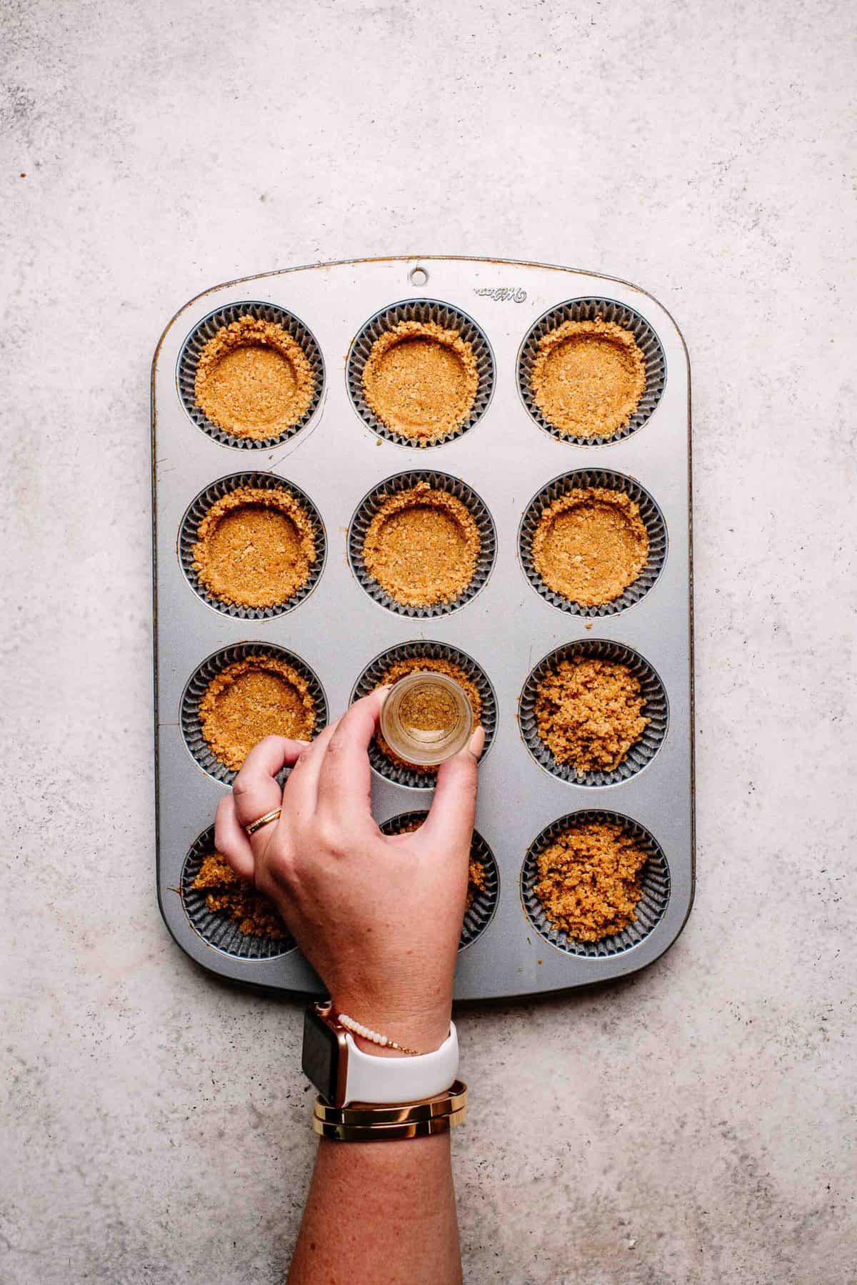 Graham cracker crust being pressed into muffin pan.