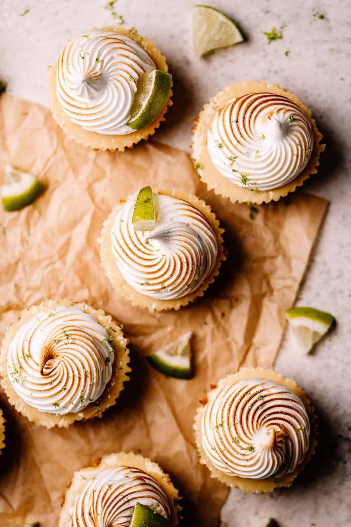 Mini key lime pies with meringue and a slice of lime on brown parchment paper.