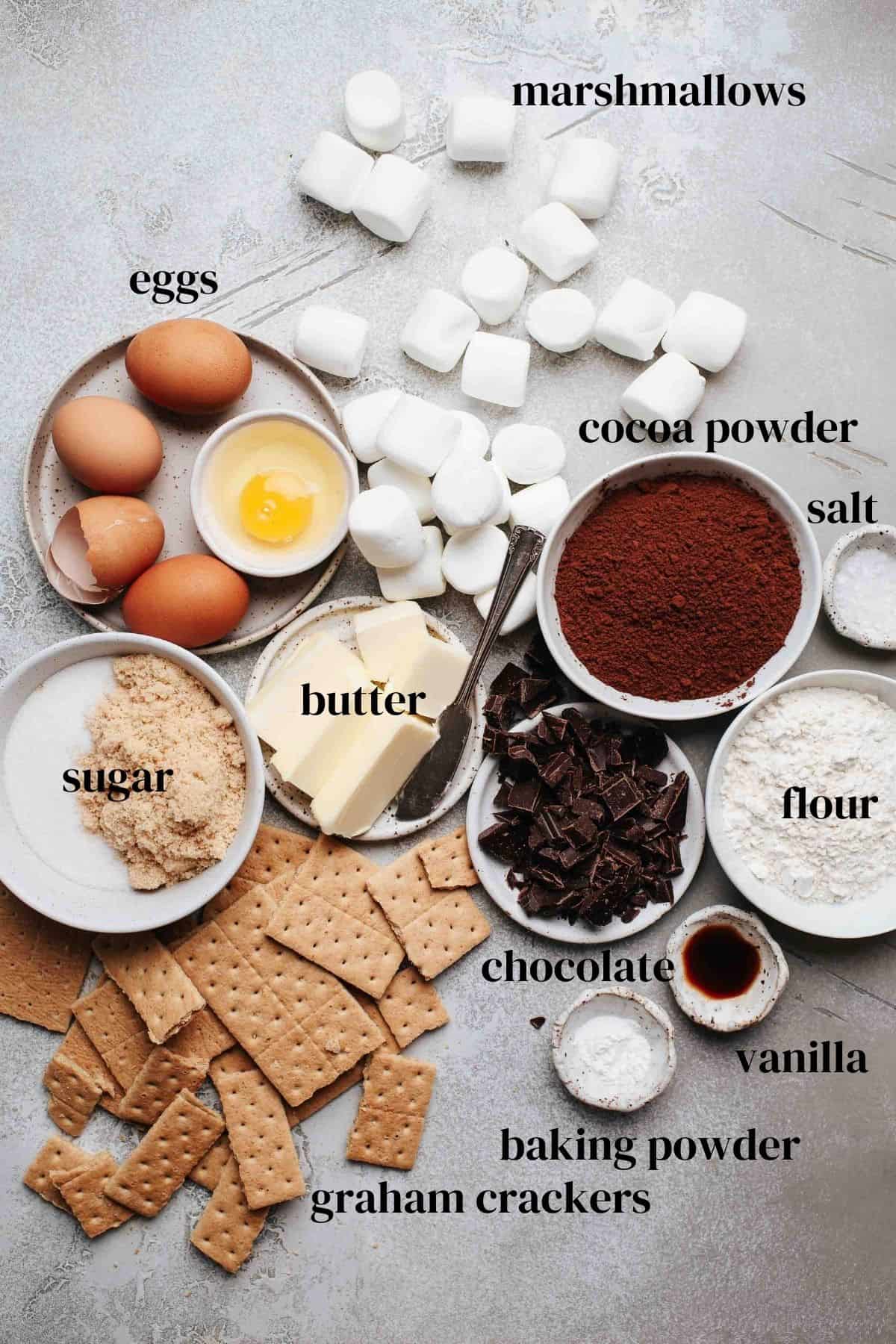 Marshmallows, eggs, sugar, butter, cocoa powder, salt, flout, chocolate, vanilla, graham crackers on a gray surface.